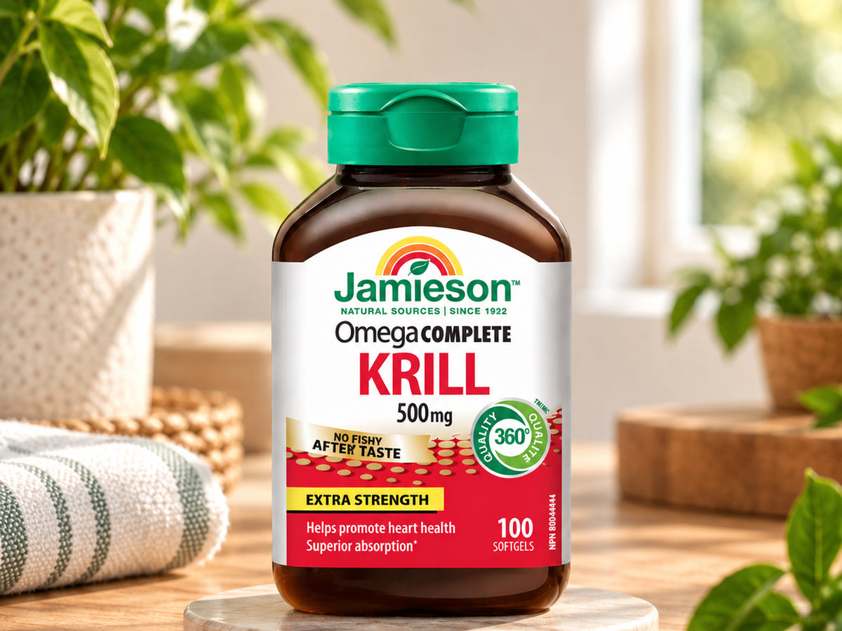 A bottle of Jamieson Omega Complete Krill 500mg dietary supplement, displayed on a wooden surface with a blurred background of potted plants and a window.