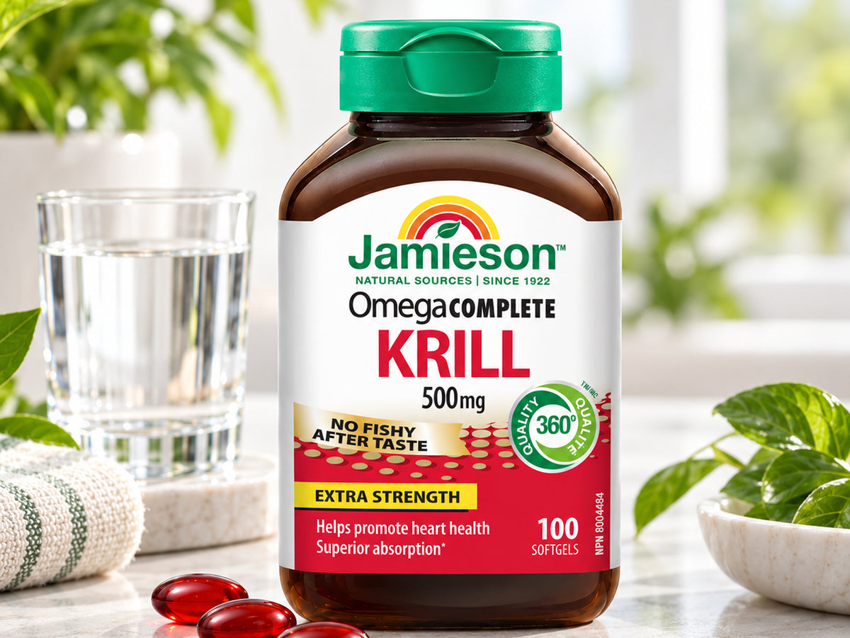 Jamieson Omega Complete Krill 500mg supplement bottle with a green cap, shown on a light marble surface with a glass of water, green leaves, and red softgel capsules in the foreground.