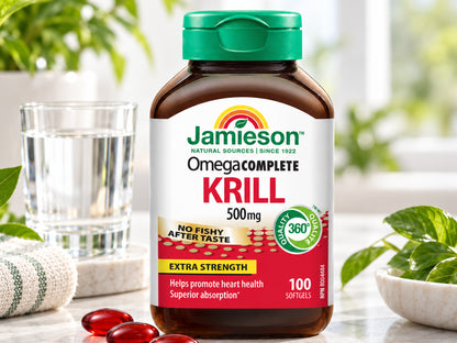 Jamieson Omega Complete Krill 500mg supplement bottle with a green cap, shown on a light marble surface with a glass of water, green leaves, and red softgel capsules in the foreground.